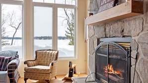 A cozy living room with a stone fireplace, a comfortable armchair, and a large window overlooking a snowy landscape. - Lakeside Memories Await at Trenanthia's Cahoots Cottage (Gravenhurst)