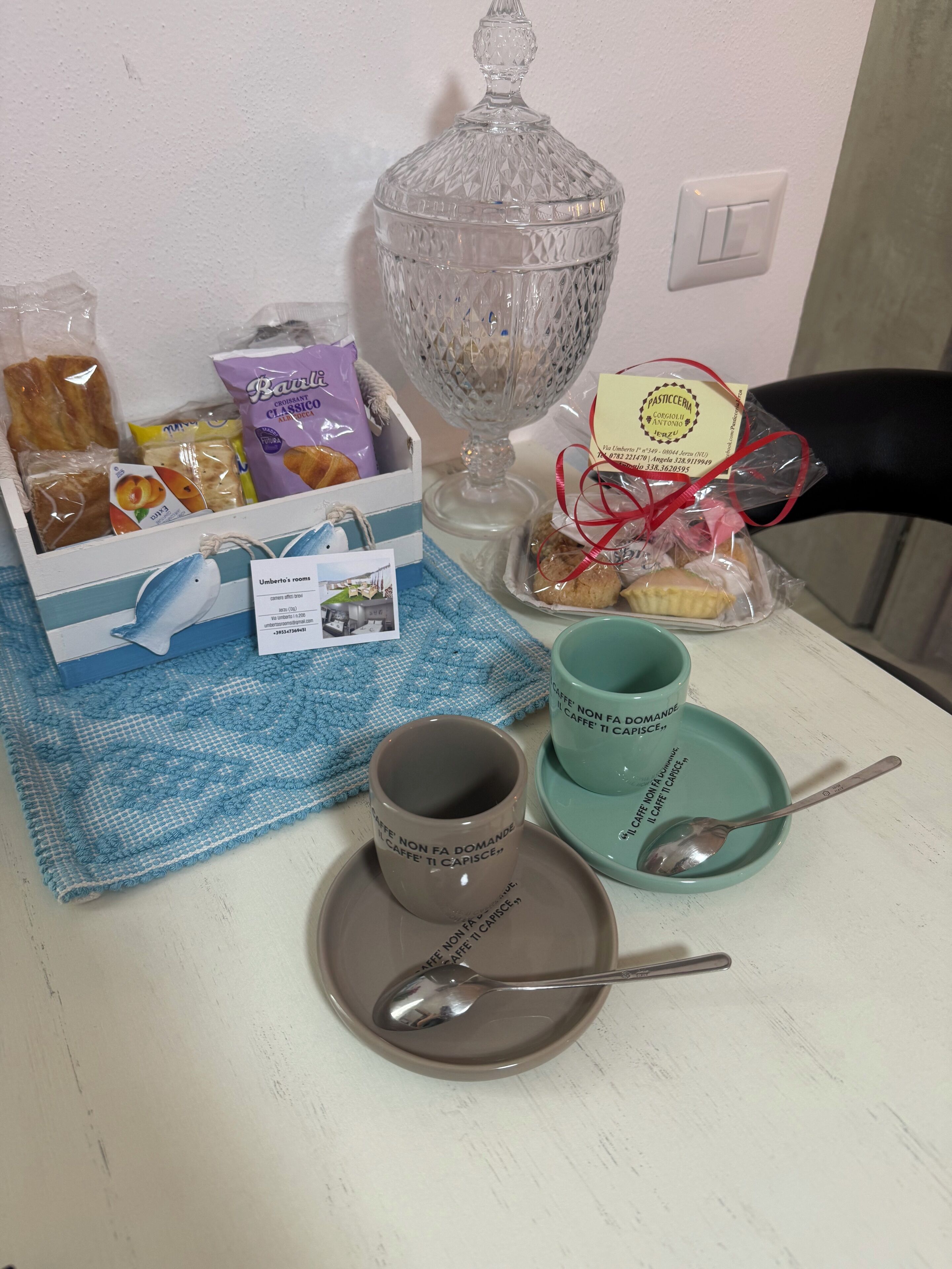 Una mesa con un decantador de vidrio, una caja de pasteles, una taza de café, una cuchara y un mantel individual.