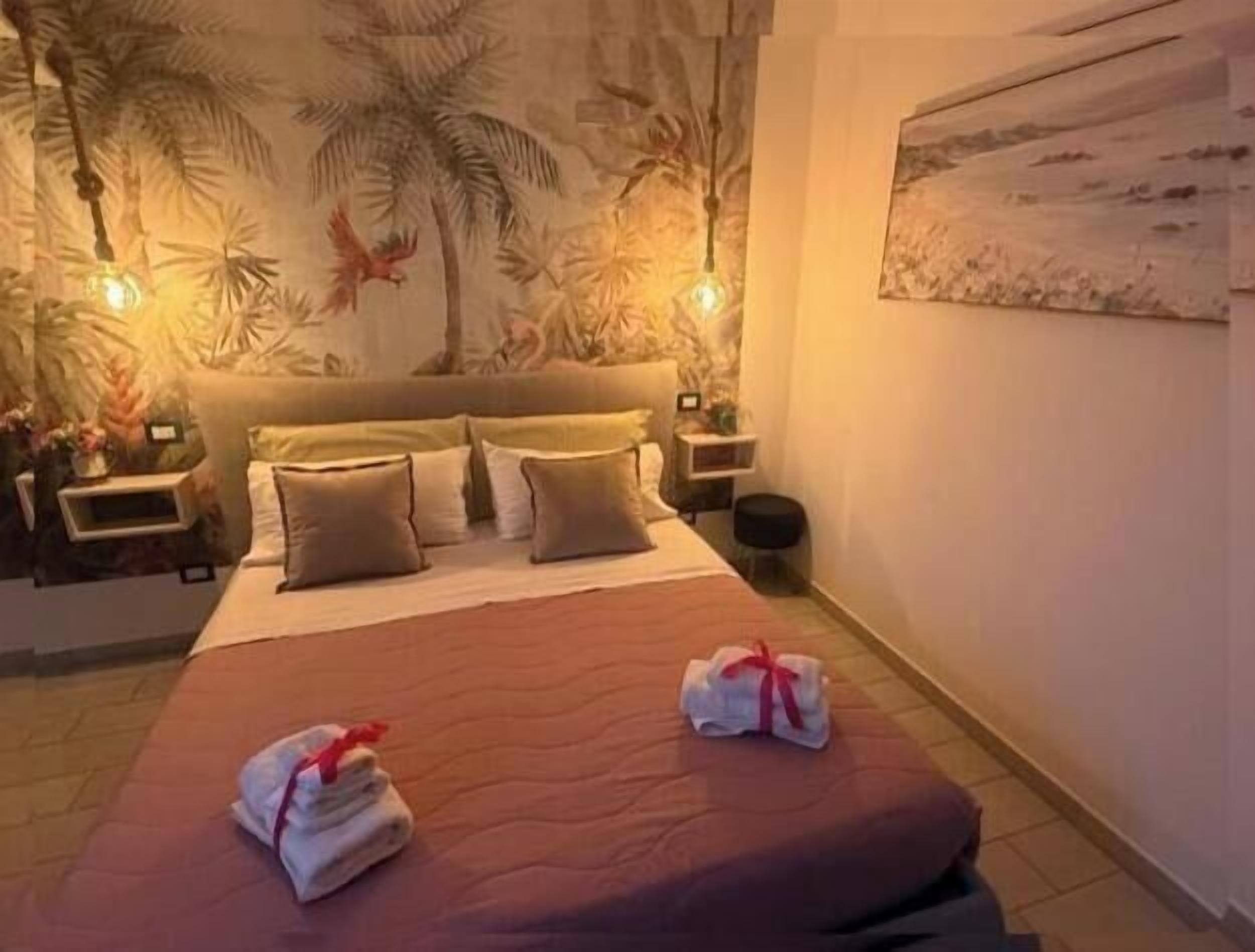 Een slaapkamer met een bed, kussens, een nachtkastje, een wandlamp en een schilderij.