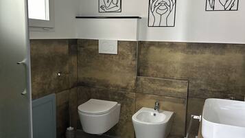 A modern bathroom with two bidets, a sink, and a mirror. There are abstract art pieces on the wall and a window above the bidets.