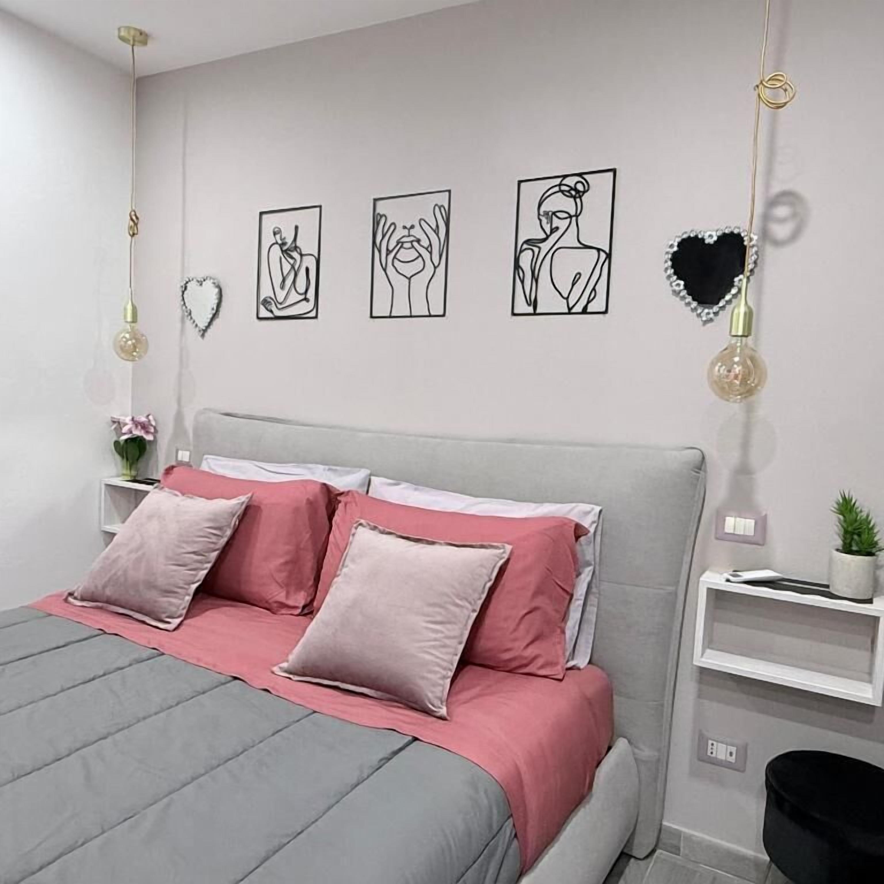 Un dormitorio moderno con cama gris, almohadas rosadas y blancas, y cabecero gris. Hay obras de arte en la pared y una planta pequeña en la mesita de noche.