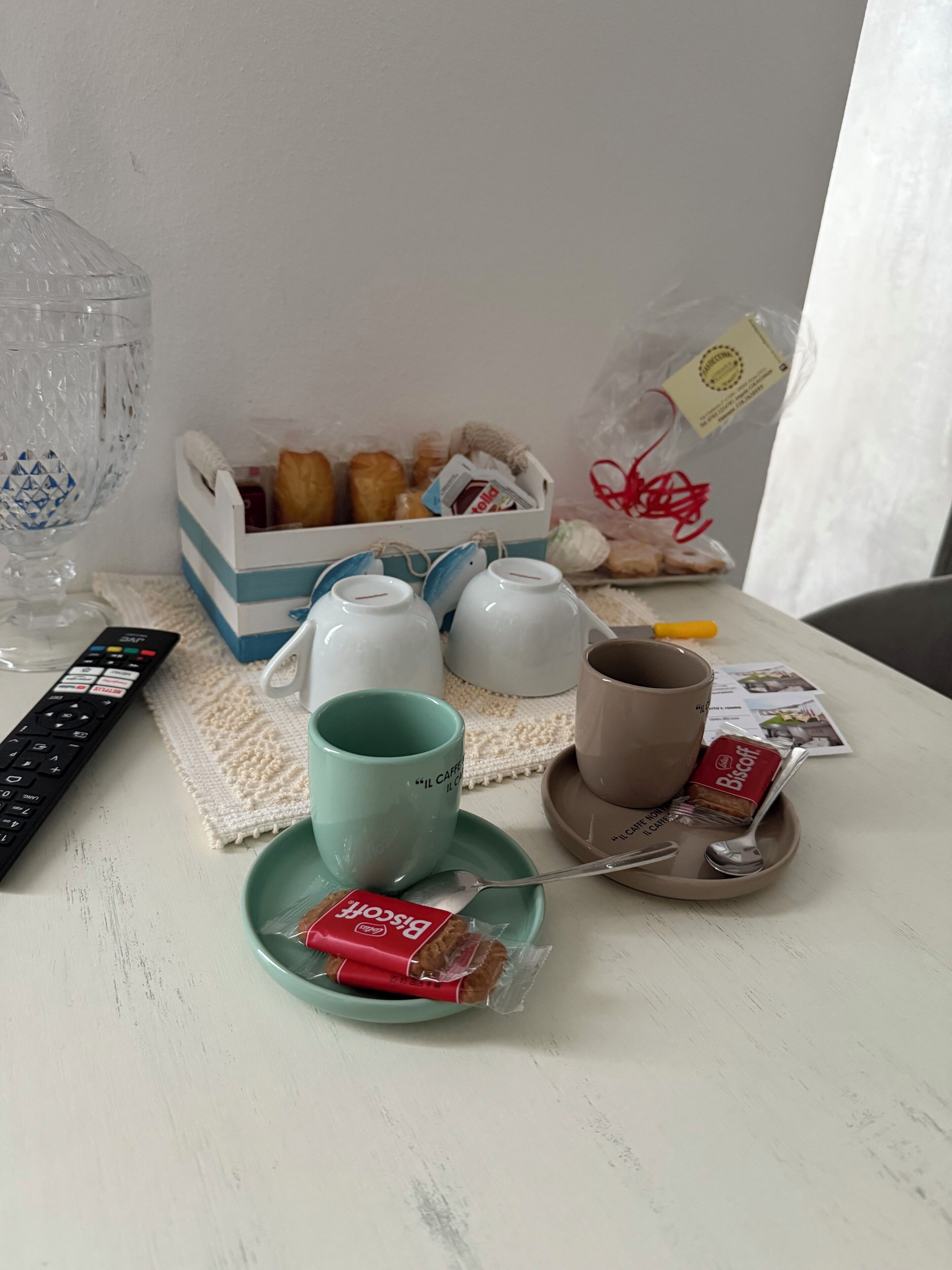 A table with a remote control, a teapot, cups, a teacup, a biscuit packet, and a box of biscuits.