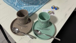 Een tafel met een glazen koepel die gebakjes bevat, een blauwe gehaakte onderzetter, twee koffiekopjes met bijpassende schotels en een lepel.