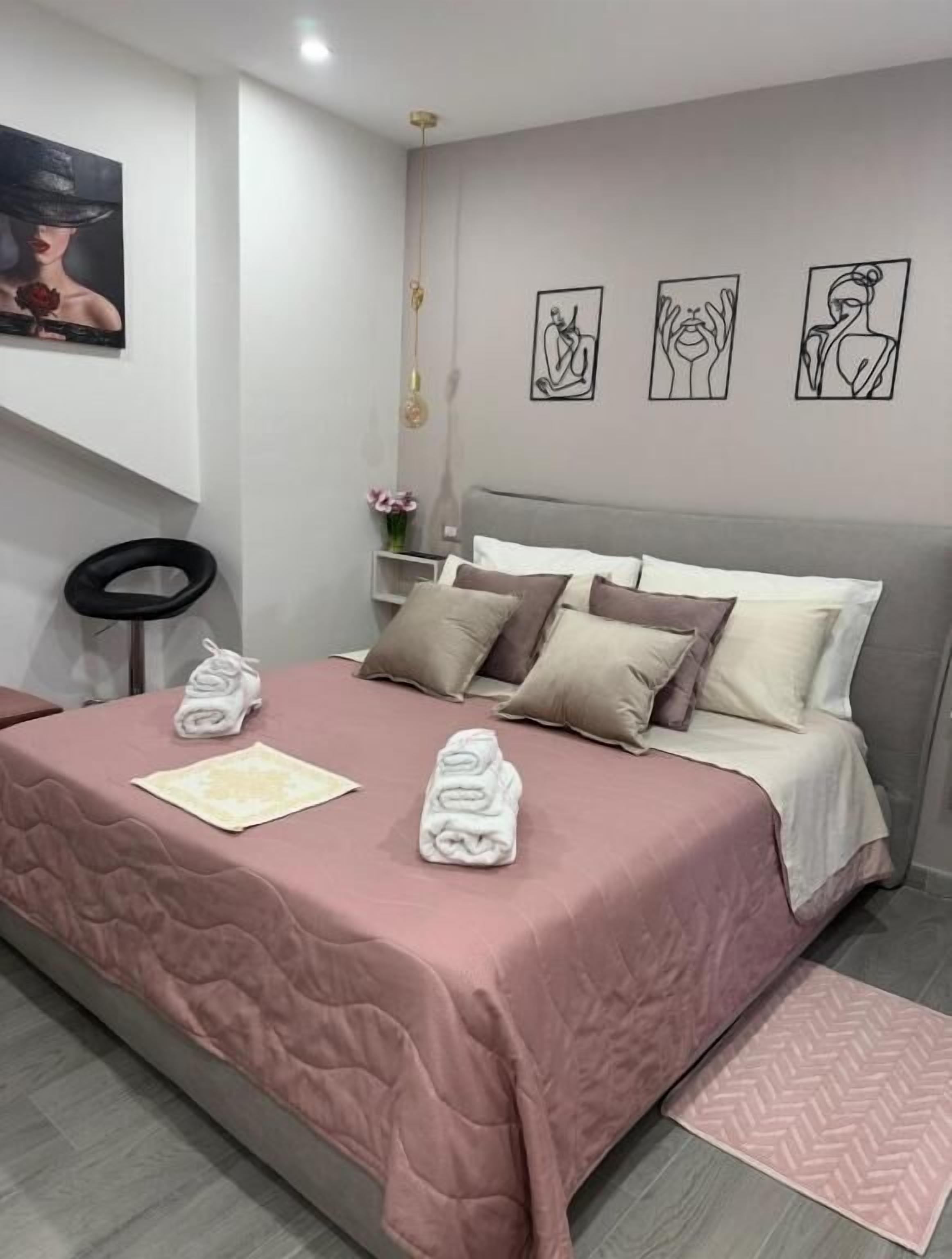 Una cama bien hecha con una colcha rosa y varios almohadones, un taburete negro circular y una mesita de noche con un florero.