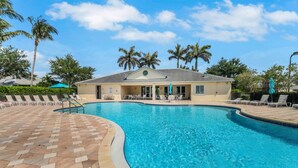 A swimming pool with lounge chairs and umbrellas, surrounded by palm trees and a building with a tiled roof. - 15041 Sandpiper Preserve Blvd #202 (Fort Myers)