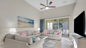 A living room with a beige sofa, a coffee table, and a TV. There is a painting on the wall and a ceiling fan. - 15041 Sandpiper Preserve Blvd #202 (Fort Myers)