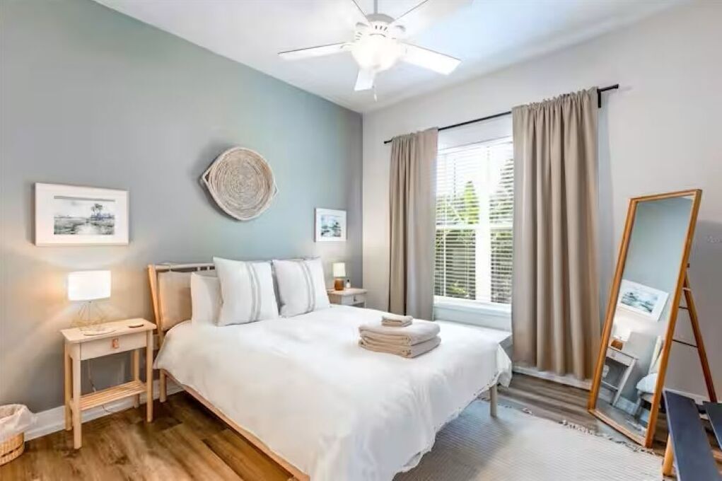 A bedroom with a bed, bedside tables, a ceiling fan, a window with curtains, and artwork on the walls.