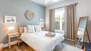 Quarto com cama, mesas de cabeceira, ventilador de teto, janela com cortinas e obras de arte nas paredes.