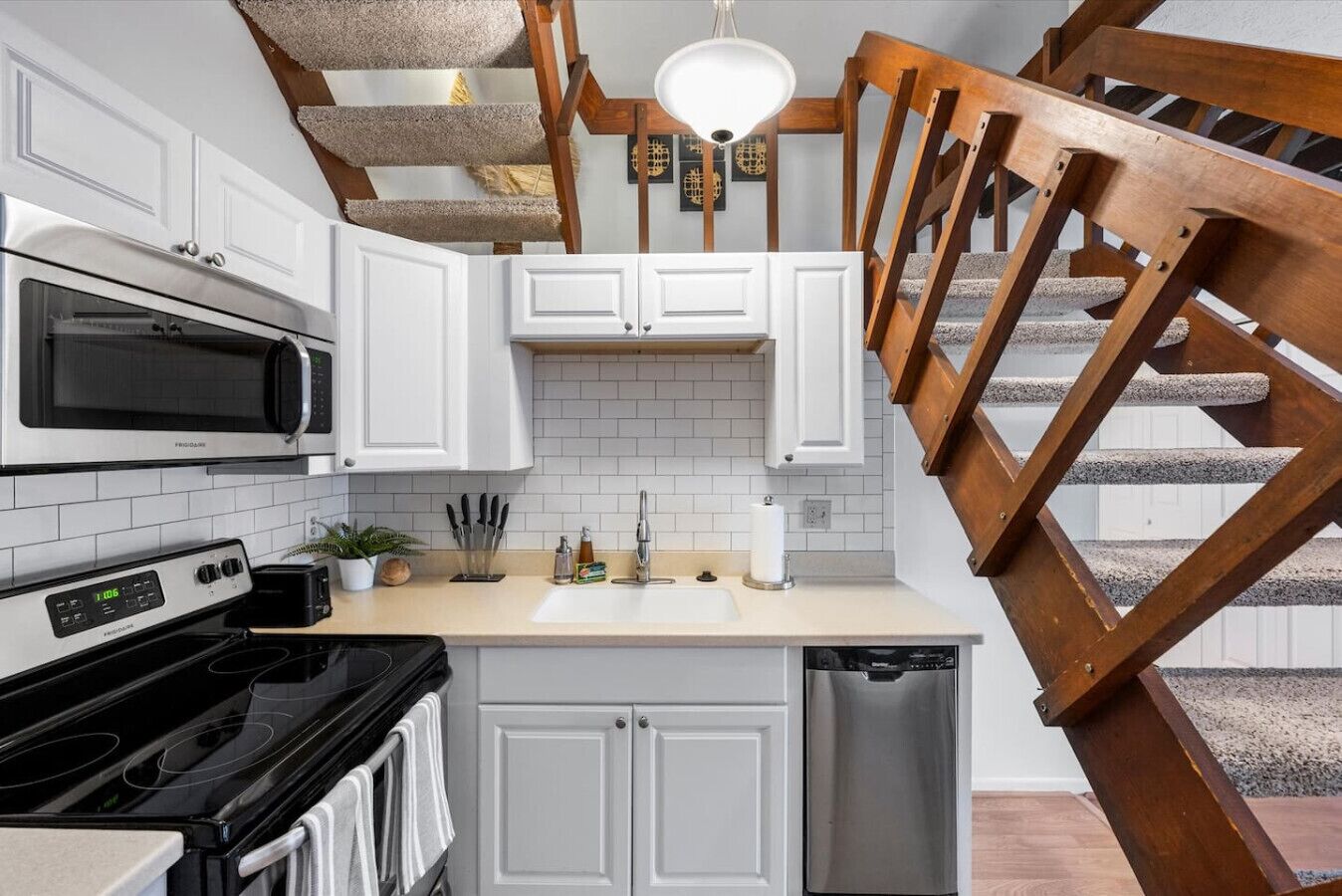 Une cuisine avec des meubles blancs, un four à micro-ondes, une cuisinière et un lave-vaisselle. On y trouve un escalier en bois avec une rampe.