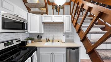 Une cuisine avec des meubles blancs, un four à micro-ondes, une cuisinière et un lave-vaisselle. On y trouve un escalier en bois avec une rampe.