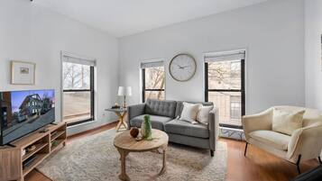 Un salon moderne avec un canapé gris, un fauteuil beige, une table basse en bois et un téléviseur posé sur un meuble en bois.