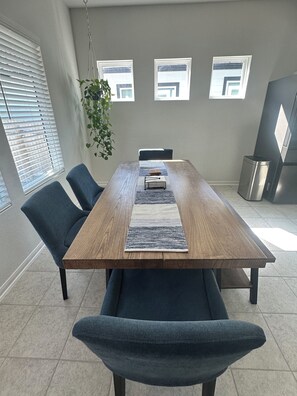 A modern dining area with a wooden table, blue chairs, a hanging plant, and a view of the city through windows. - Play and Stay Spacious Luxe Lounge (Hutto)