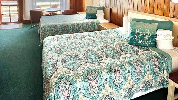 A room with two beds, a table, and a chair. The room has wooden paneling on the walls and a view of the ocean through the windows. - Sea Foam Motel (Nags Head)