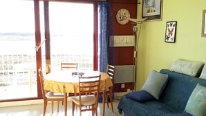 Une pièce comprenant une table à manger et des chaises, un canapé, une horloge et une vue sur l’extérieur, accessible par une porte coulissante.