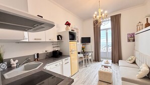 A modern kitchen with a built-in stove, a dining area with a table and chairs, a sofa, and a large window with curtains. - Studio for 2 with Wi-Fi, residence with pool (Les Sables-d'Olonne)