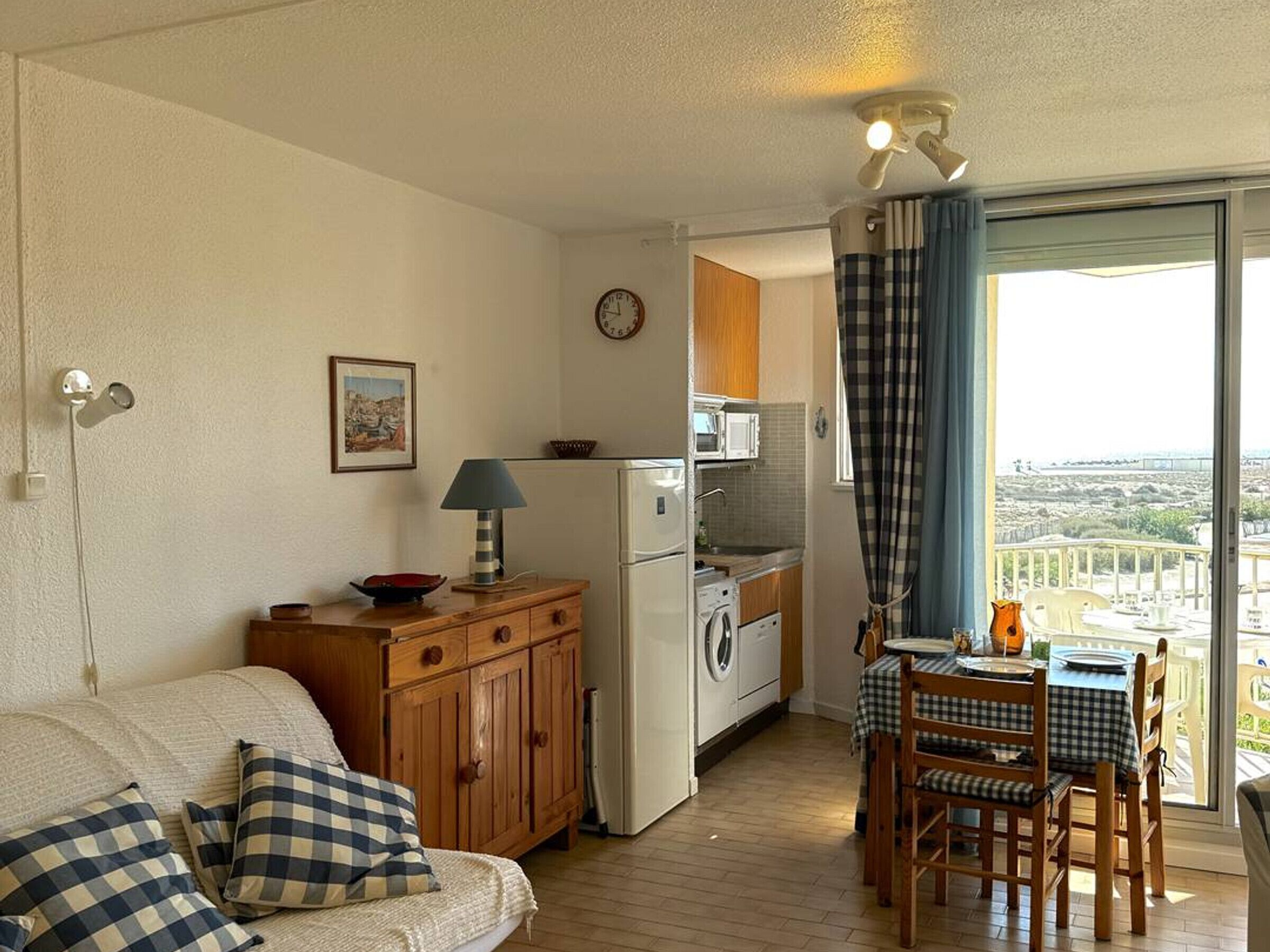 Un espace de vie compact comprenant un coin cuisine, un coin repas avec une table et des chaises, un canapé avec des coussins à carreaux et une vue dégagée sur l’extérieur grâce à une grande fenêtre.