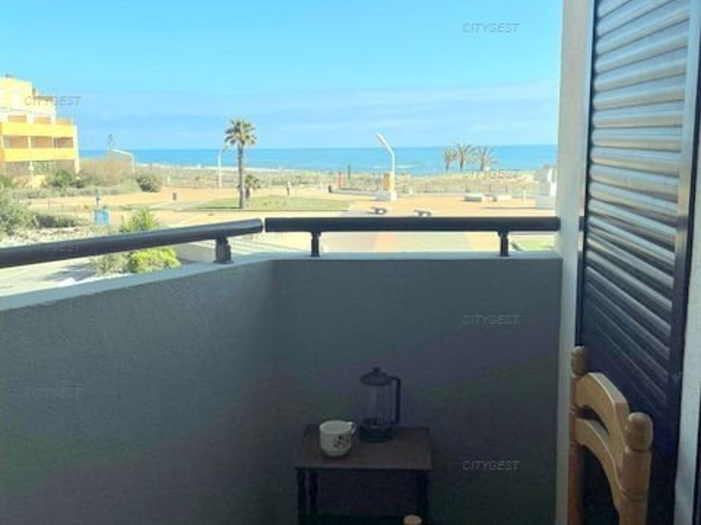 En balkong med utsikt över stranden och havet. Där står ett litet bord med en kopp och en behållare på.