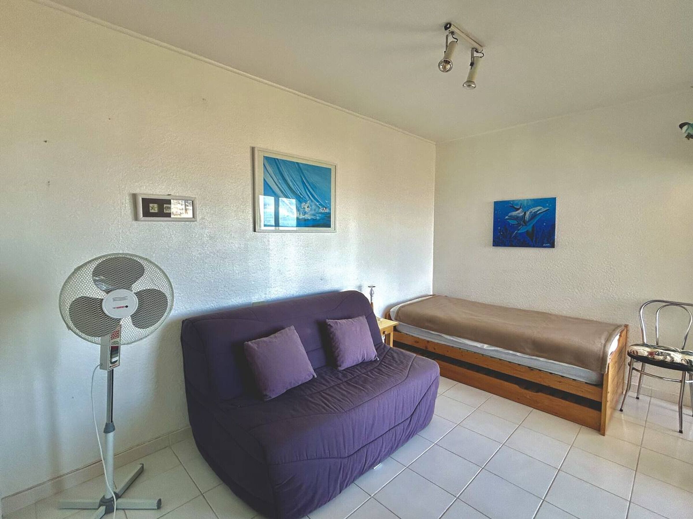 Una habitación con un sofá morado, una cama individual, un ventilador de pie y dos lámparas de pared.