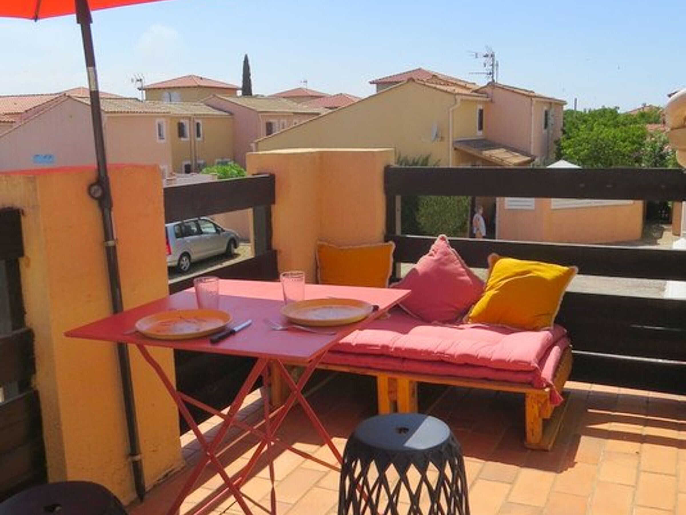 Een balkon met een rode tafel, twee stoelen, een roze bank en een zwarte kruk.