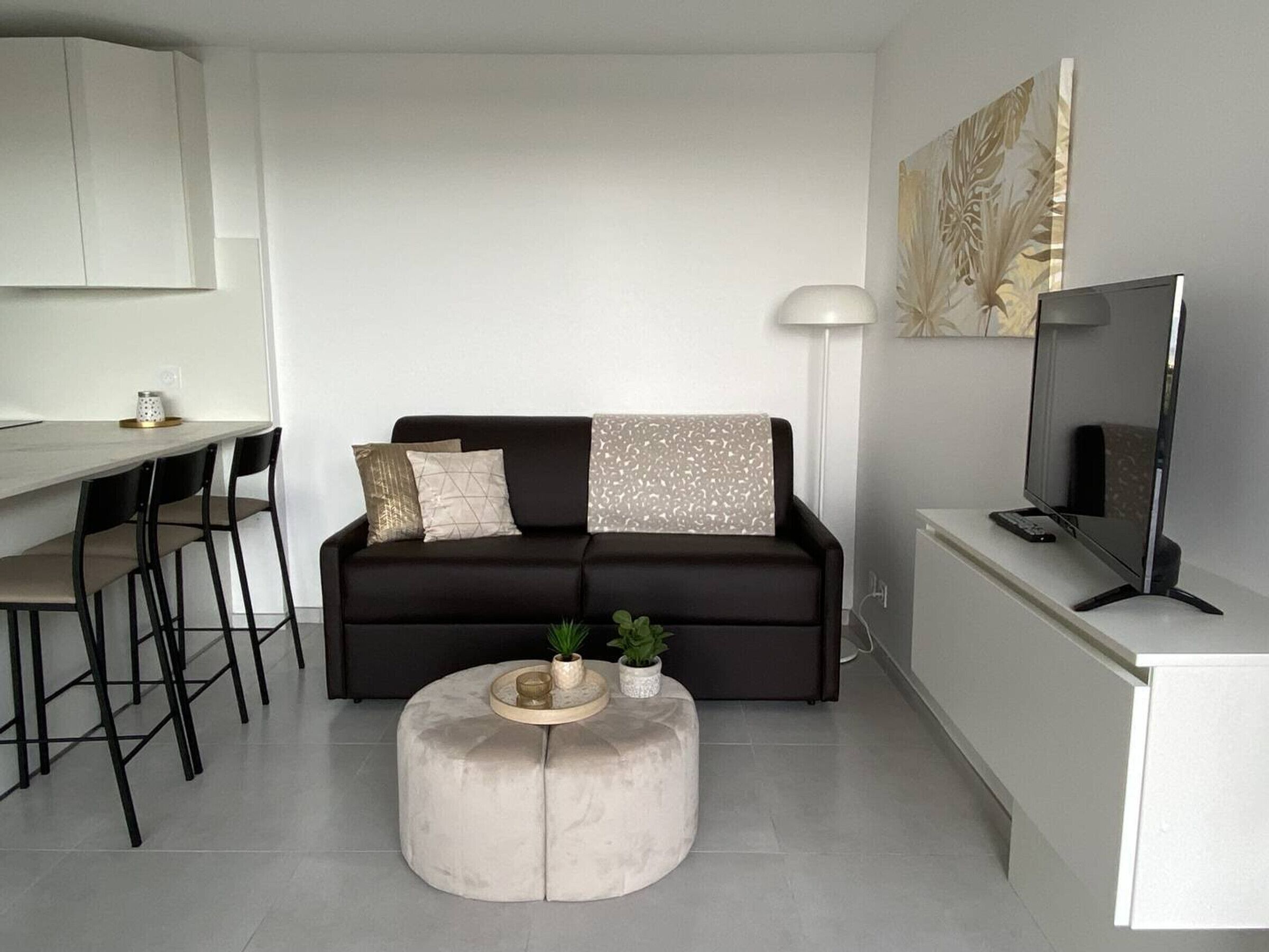 A modern living room with a dark brown sofa, a round ottoman, a flat-screen TV, and a dining area with a white table and black chairs.