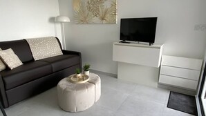 Ein modernes Wohnzimmer mit einem dunklen Sofa, einem runden Hocker, einem Flachbildfernseher und einem schlichten, weißen Schreibtisch.