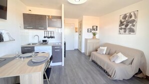 A modern kitchen with grey cabinets, a dining table with chairs, and a sofa in a living area. - Cozy studio beach access, balcony and parking (Le Barcarès)