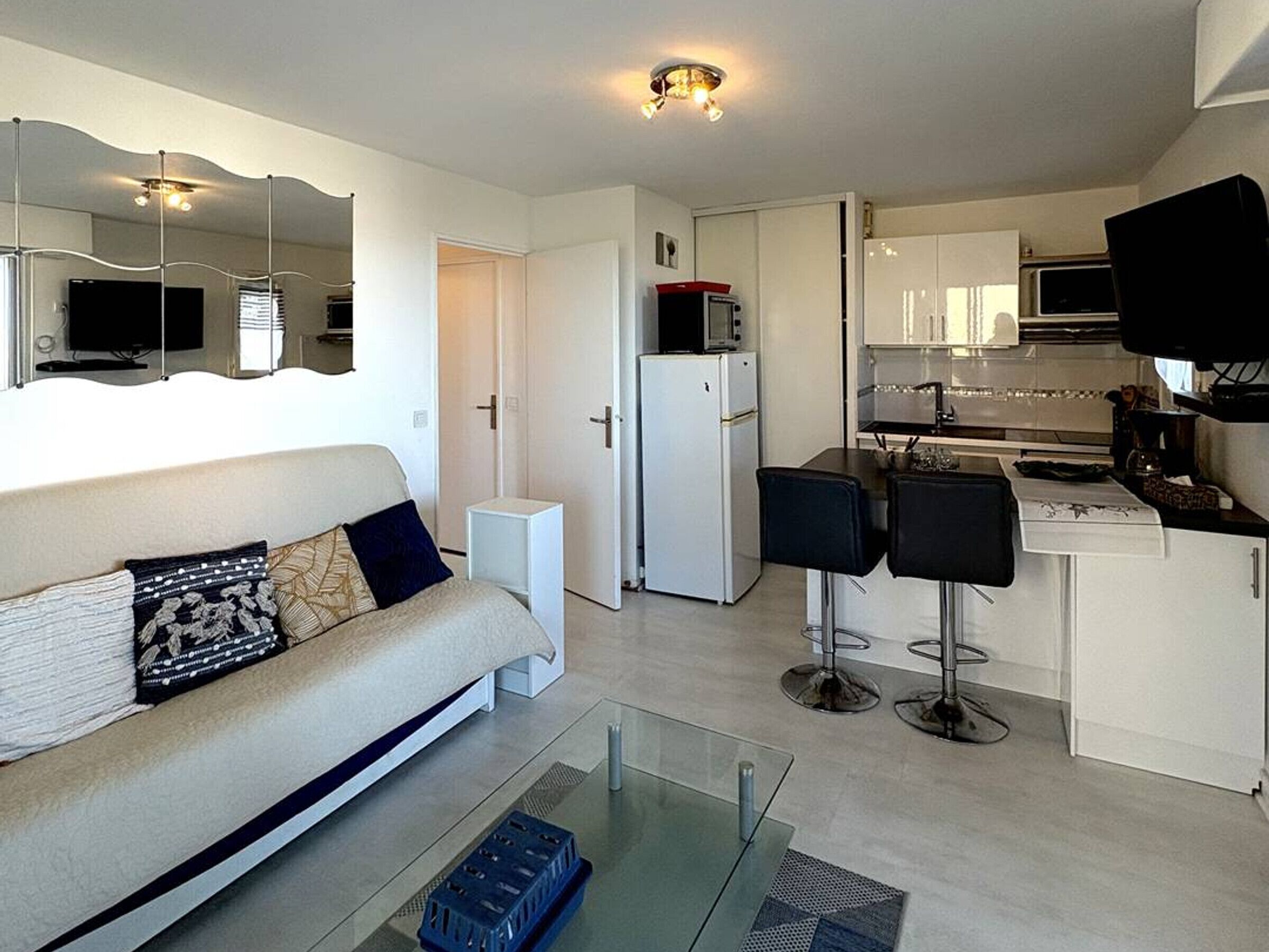 A modern living room with a sofa, a glass coffee table, and a kitchen area with a microwave, refrigerator, and bar stools.
