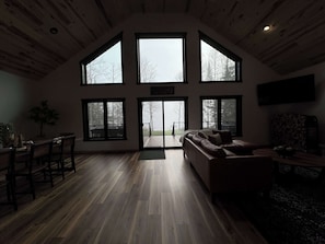 Ein geräumiges Wohnzimmer mit Holzboden, einer großen Couch und einem Essbereich mit Stühlen und Tisch. Mehrere große Fenster sorgen für natürliches Licht und Blick ins Freie.