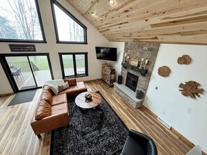 Ein modernes Wohnzimmer mit einer Ledersofa, einem runden Couchtisch, einem Steinkamin und großen Fenstern mit Blick auf eine Terrasse.