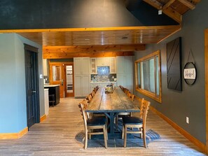 A spacious dining area with a long wooden table and chairs, a kitchen in the background, and a large window. - Larrabee Lake Retreat - The Lodge & Cottage (New Auburn)