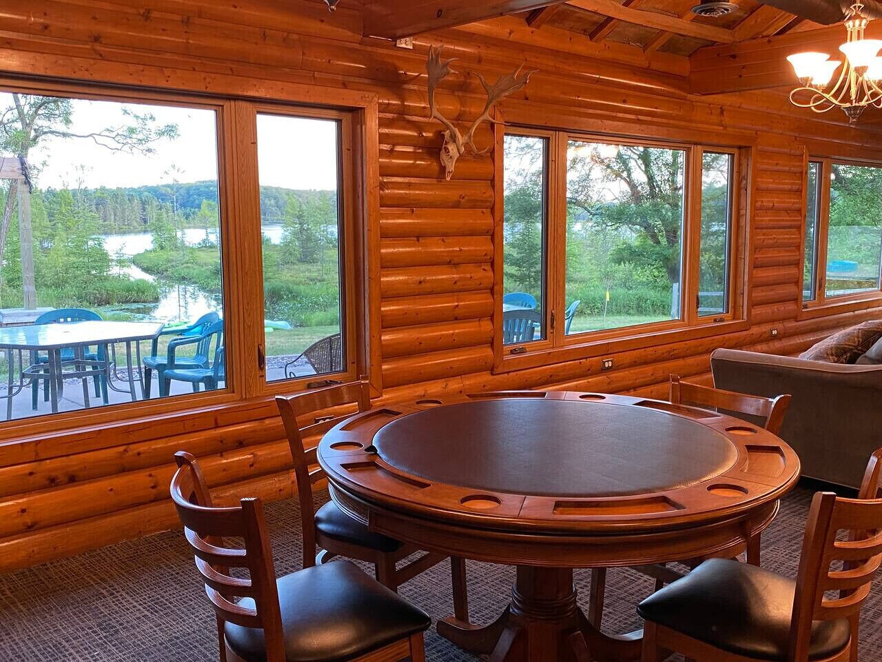 L’intérieur d’une cabane en rondins, avec une table et des chaises en bois rond, de grandes fenêtres donnant sur un lac et une forêt, et un lustre suspendu au plafond.