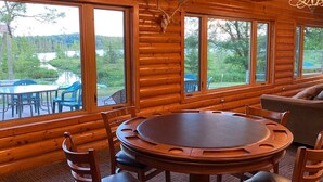 A log cabin interior with a round wooden table and chairs, large windows offering a view of a lake and forest, and a chandelier hanging from the ceiling.