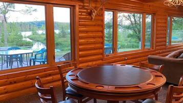 L’intérieur d’une cabane en rondins, avec une table et des chaises en bois rond, de grandes fenêtres donnant sur un lac et une forêt, et un lustre suspendu au plafond.