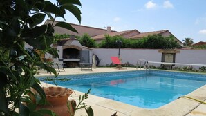 Ein Schwimmbecken mit Fliesenbordstein, umgeben von einer Terrasse mit Grill und Gartenmöbeln.