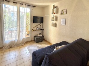 Sala de estar com sofá escuro, TV de tela plana na parede, um pequeno armário e janela com cortinas leves.