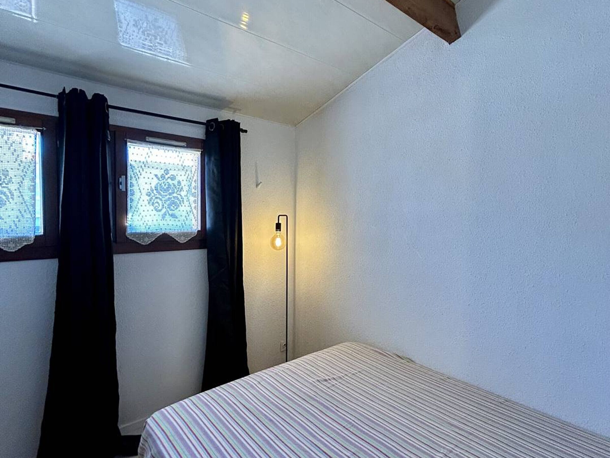 A small, single-bed room with a plain white wall, a window with lace curtains, and a floor lamp.