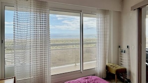 A bedroom with a large window, a bed, a small stool, and a radiator.