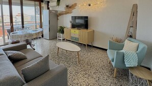 A modern living room with a sofa, a TV on a wooden cabinet, a dining table with chairs, and a large window offering a city view. - Apartment 4 people with terrace, AC, parking, 200m from beach (Le Barcarès)