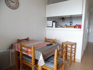 Ein kleiner, gemütlicher Raum mit einem Holztisch und Stühlen, einem Bett mit gestreifter Bettwäsche, einer Uhr an der Wand und einer Küchenzeile mit einer Mikrowelle und kleinen Geräten.