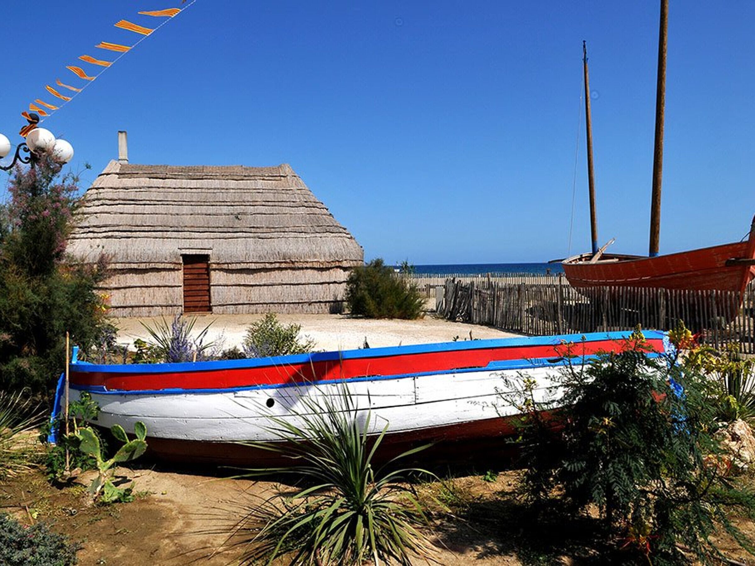 Una casa con techo de paja, un bote pequeño y una valla de madera junto a la playa.