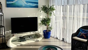 A modern living room with a curved glass coffee table, a flat-screen TV, a potted plant, and a wicker chair. - Modern apartment with AC 100 m from the beach for 6 pers (Le Barcarès)