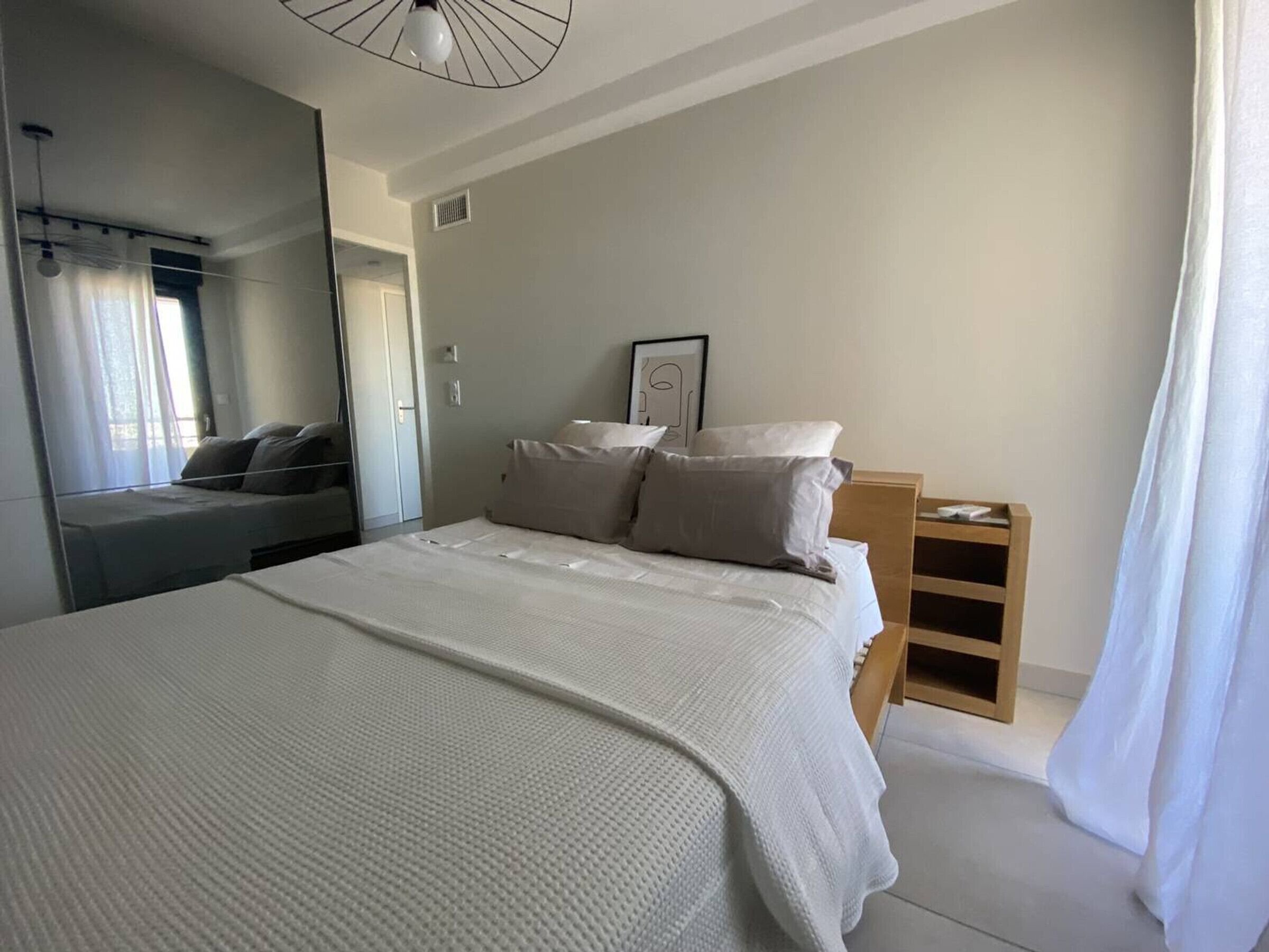A modern bedroom with a large bed, a bedside table, a mirror, and a window with curtains.