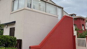 En bostadsbyggnad med en beige fasad, stora fönster med vita persienner och en röd trappa som leder till entrén.