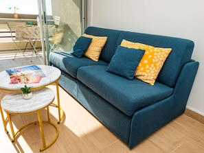 Un canapé bleu agrémenté de coussins à motifs floraux jaunes, une petite table ronde avec un plateau en marbre blanc et une plante en pot.