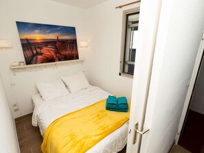 Une chambre à coucher avec un lit, un tableau représentant un coucher de soleil, une fenêtre et une porte.