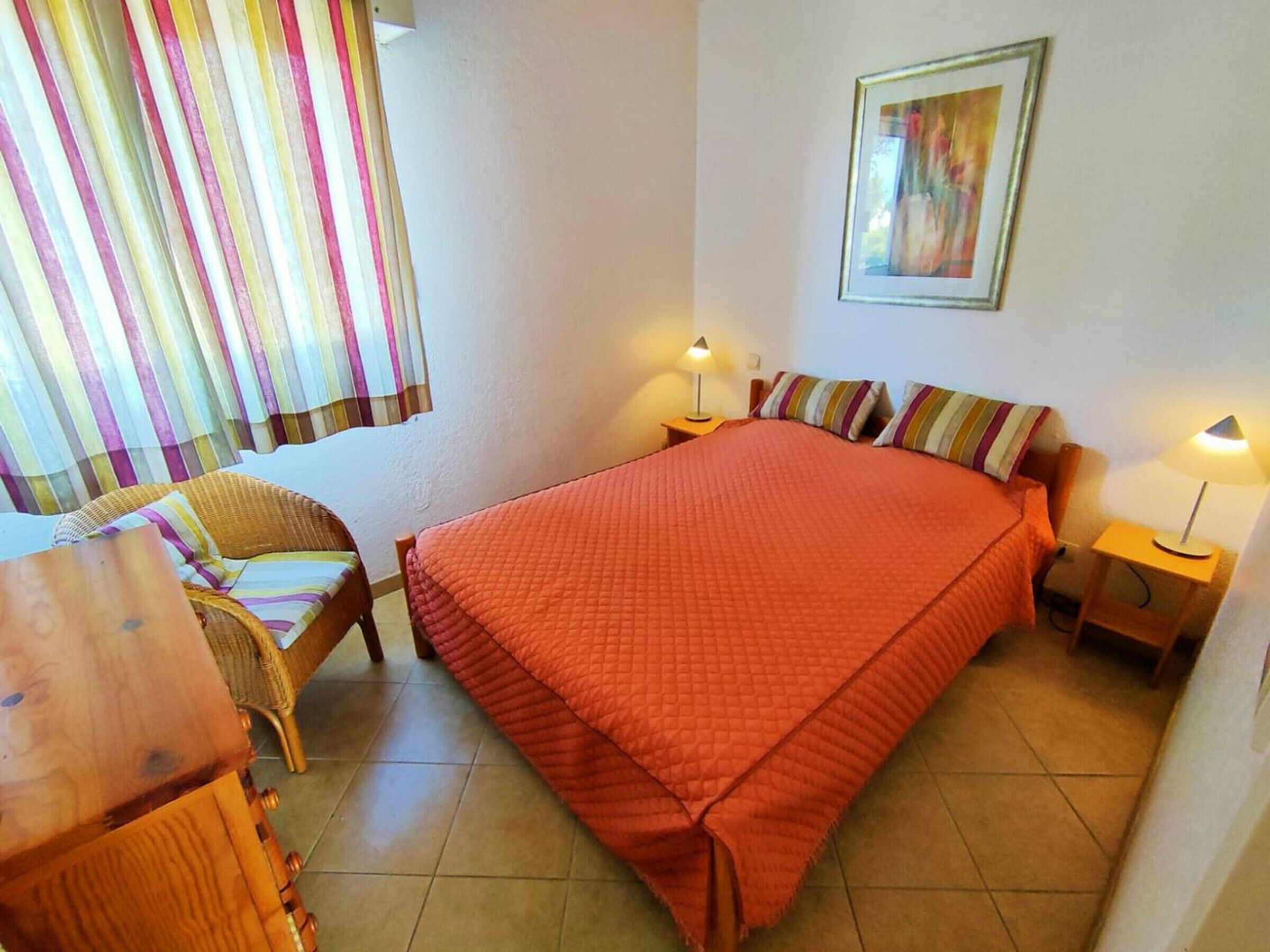 A bedroom with a bed, two bedside tables with lamps, a chair, a window with curtains, and a framed picture on the wall.