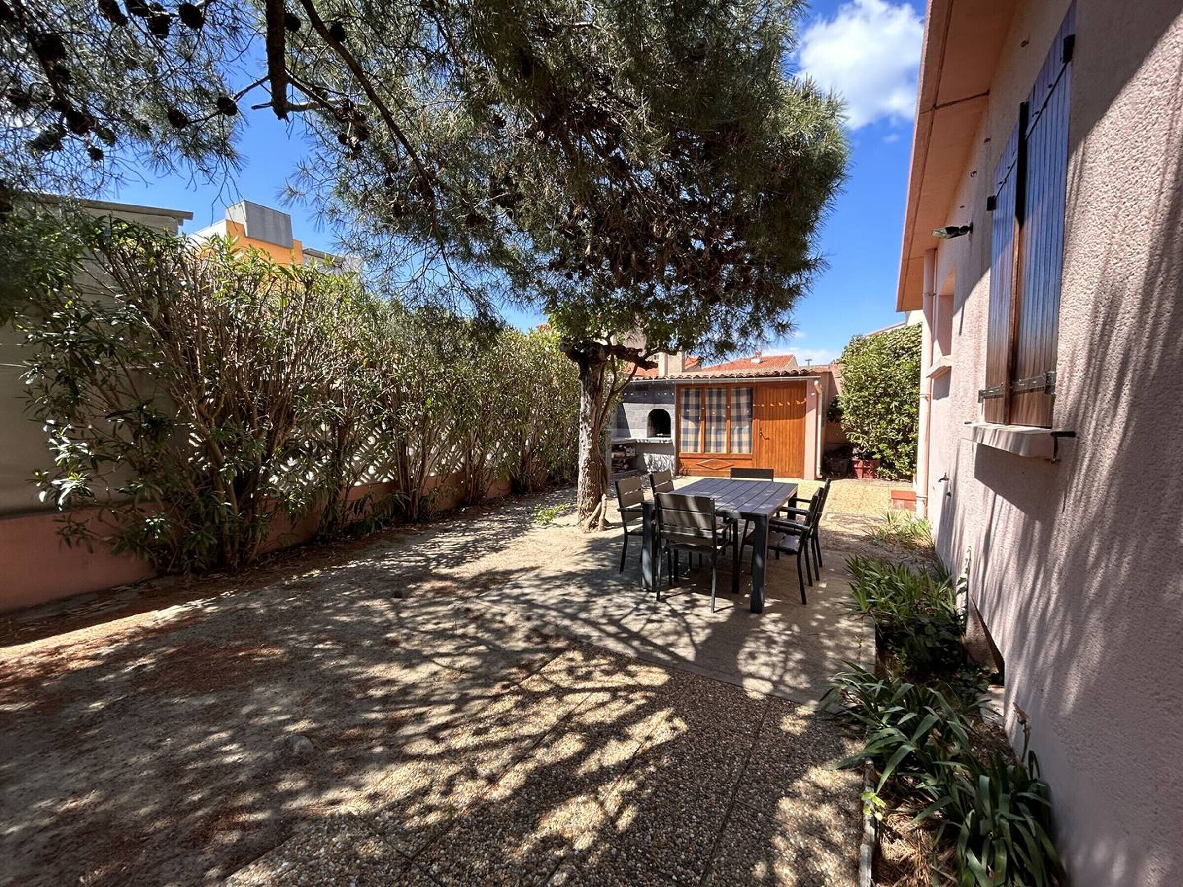A patio area with a table and chairs, surrounded by dense foliage and a building with a tiled roof.