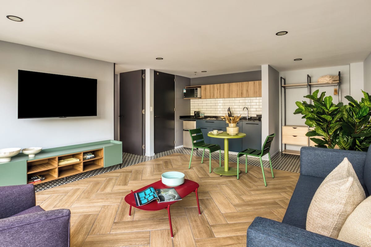 A modern living room with a flat-screen TV, a sofa, a red coffee table, and a dining area with a small table and chairs.