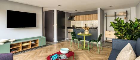 A modern living room with a flat-screen TV, a sofa, a red coffee table, and a dining area with a small table and chairs.
