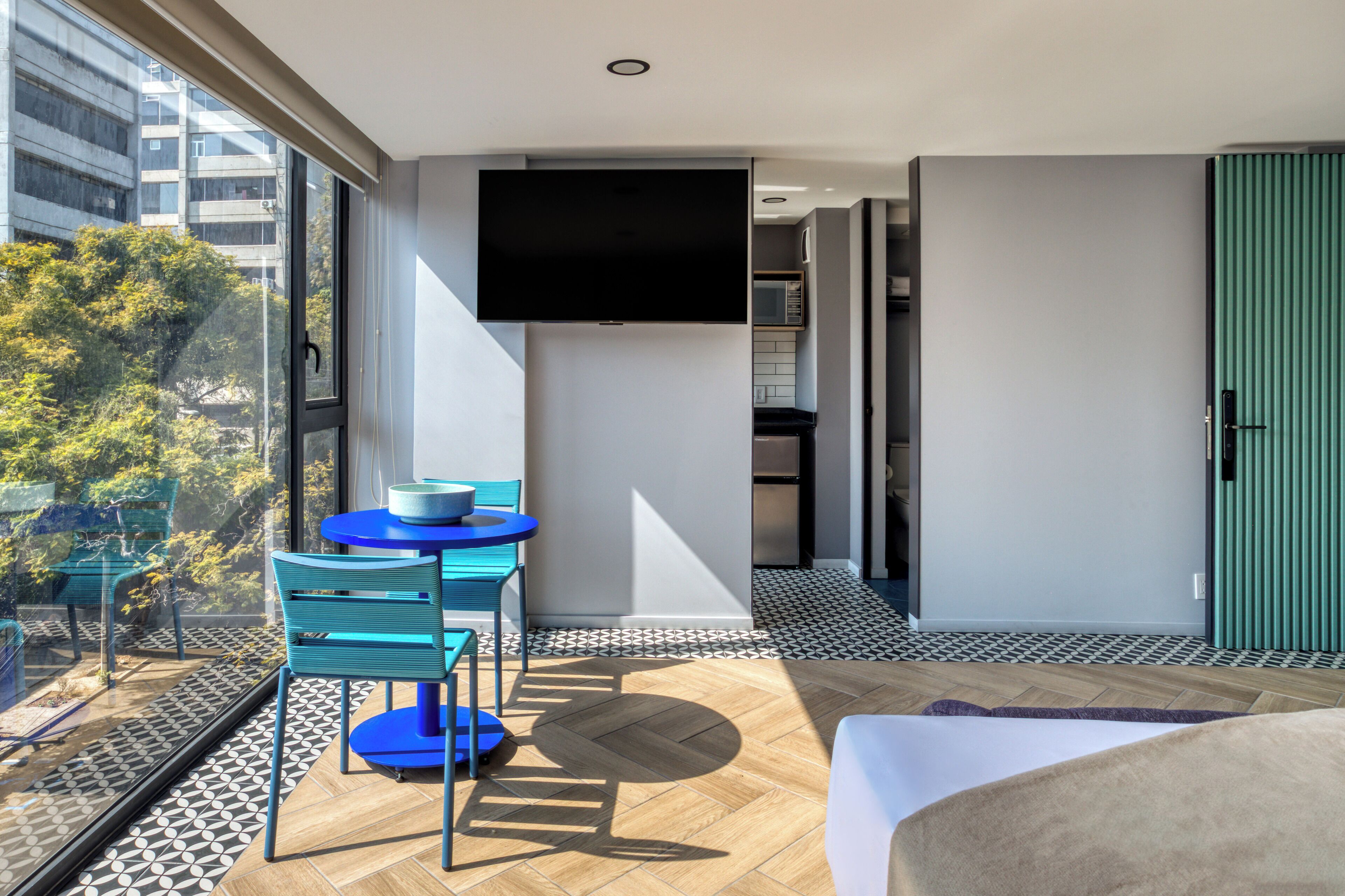 A modern hotel room with a large window, a flat-screen TV, a small table with chairs, and a view of the city.